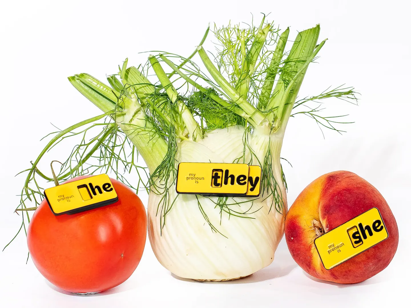 A tomato wearing a “he” pronoun pin, fennel wearing a “they” pronoun pin, and a peach wearing a “she” pronoun pin